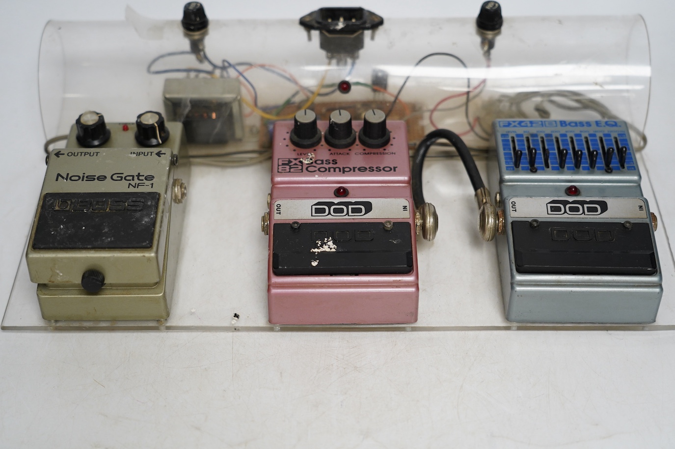 A bass guitar pedal board including; a DOD FX42 B bass EQ pedal, a DOD FX82 bass compressor and a Boss noise gate NF-1 pedal, mounted on a clear Perspex frame. Condition fair, some wear from use evident.